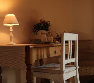 Psací stůl s ubrusem, bílá zrenovovaná židle, květiny na stole, rozsvícená lampička, v dálce hnědé křeslo.