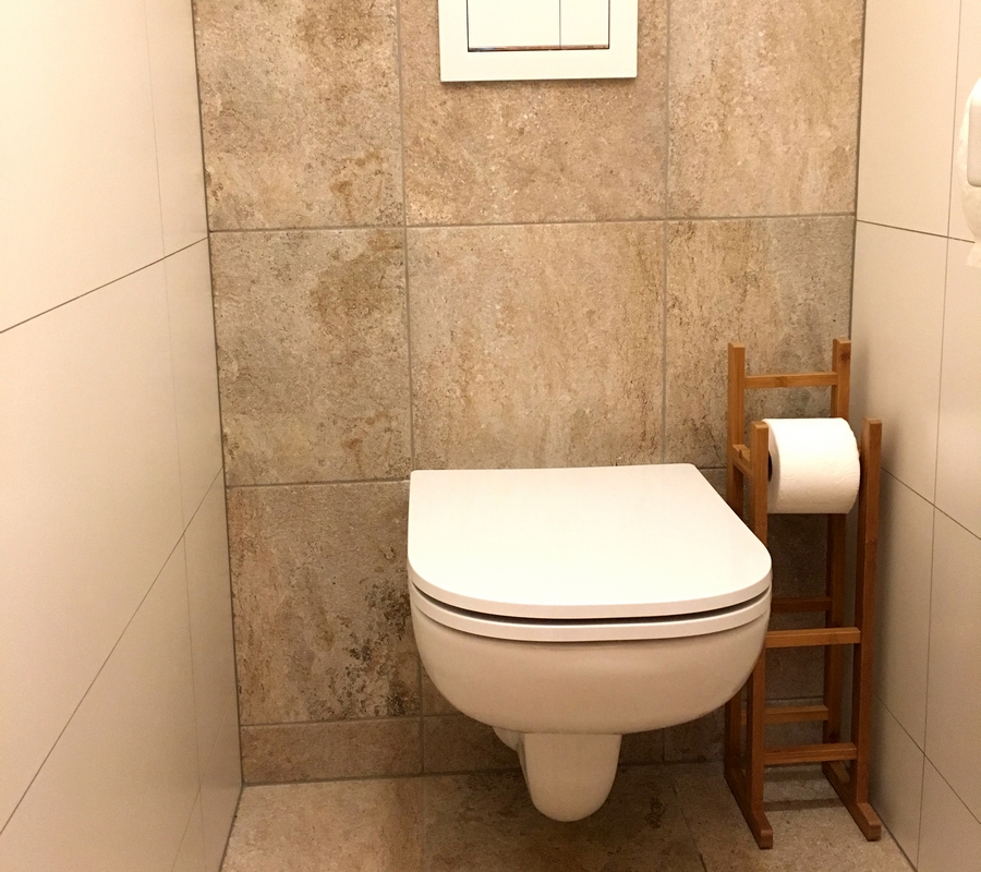 Bílý záchod, dřevěný stojan na toaletní papír, bílé a šedivé kachličky.