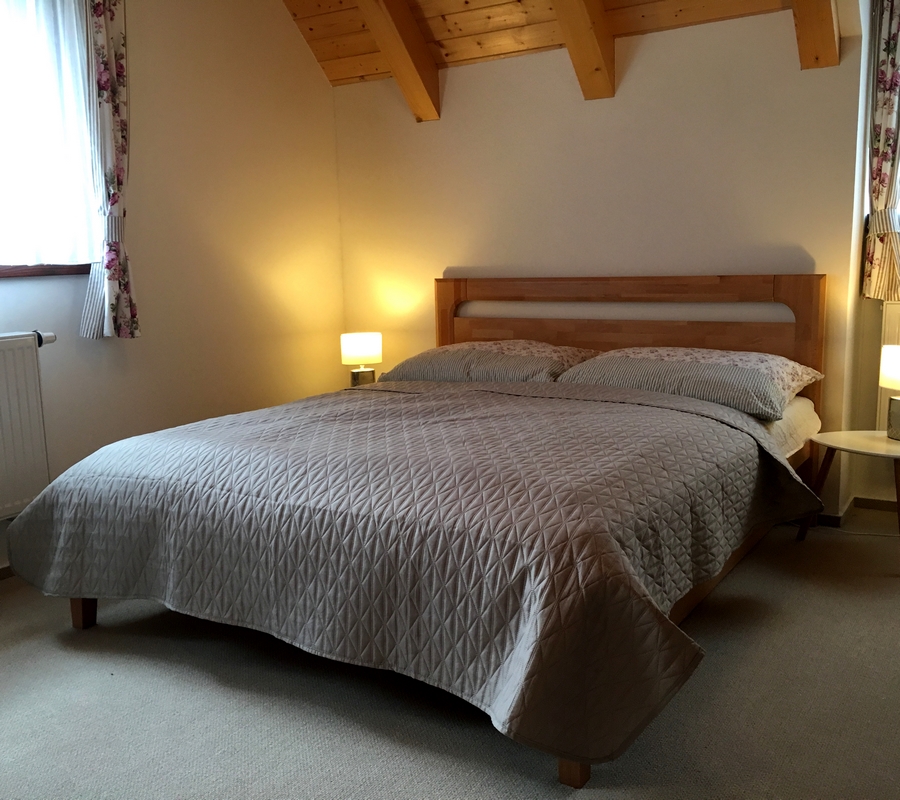Dřevěná manželská postel s šedivohnědým povlečením, noční stolky s lampičkami, květinové závěsy a trámy.