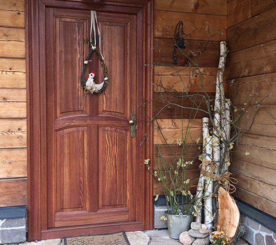 Vchodové dveře s velikonoční výzdobou, dřevěné trámy, břízy, slepička na dveřích, květiny, dekorace.