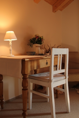Psací stůl s ubrusem, bílá zrenovovaná židle, květiny na stole, rozsvícená lampička, v dálce hnědé křeslo.