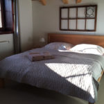 Dřevěná postel s béžovým přehozem a s béžovými ručníky, dřevěné okno jako dekorace na stěně, okrasná lampa.