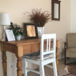 Dřevěný psací stůl, na něm položené zrcadlo, bílá lampička, bílá váza se sušenými květinami, živá dekorace, bílá dřevěná židle a v dálce hnědé zrcadlo, starožitné křeslo a kožešina na zemi.